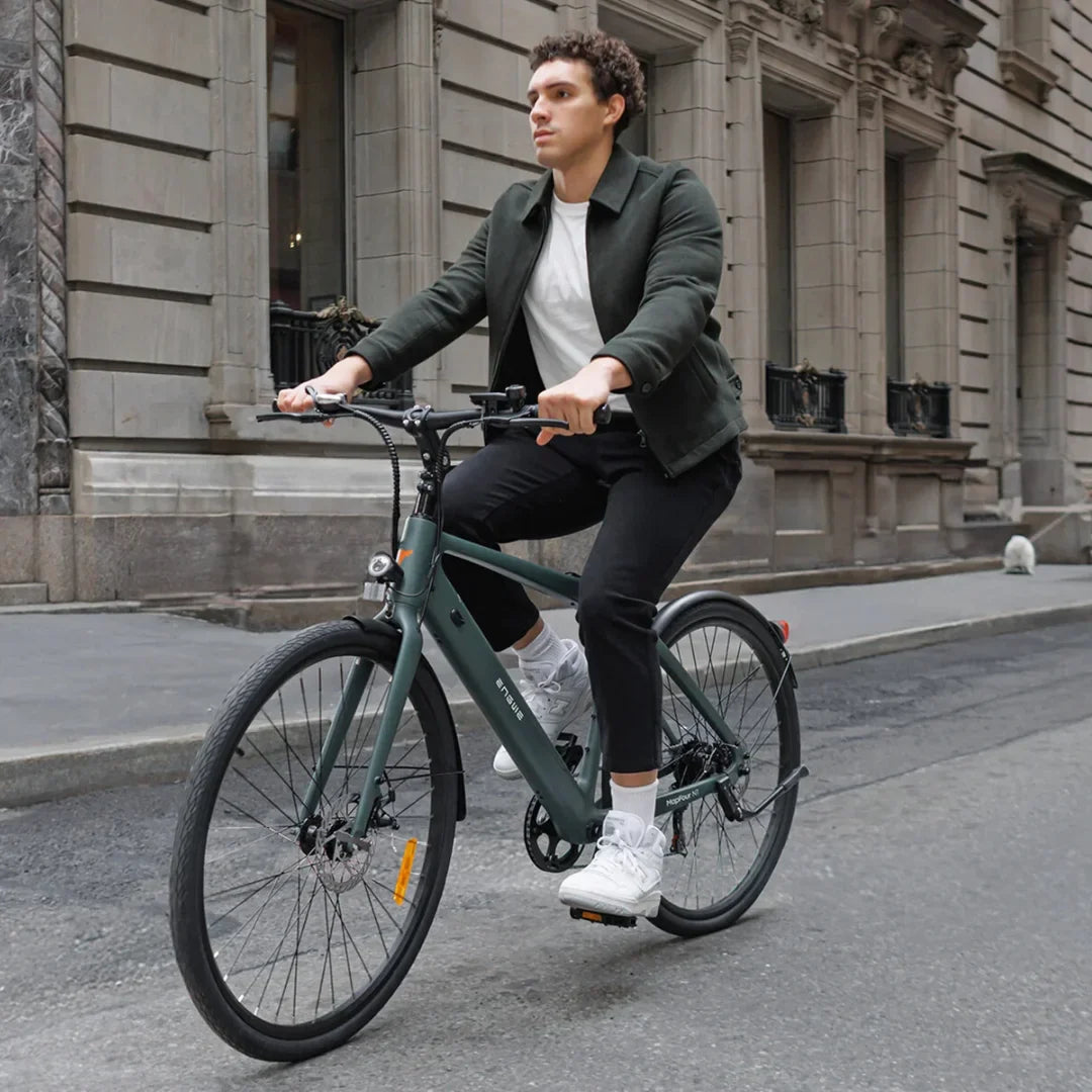Izdelek je električno kolo znamke Engwe, model Mapfour N1 Air. Na sliki je moški, ki se vozi na tem kolesu po mestni ulici. Kolo ima moderno zasnovo, z značilnimi širokimi pnevmatikami in elegantnim okvirjem v temno zeleni barvi. Moški nosi športno obleko, kar nakazuje na sproščeno vožnjo v urbanem okolju. V ozadju so vidne mestne zgradbe, kar dodatno poudarja mestno atmosfero.