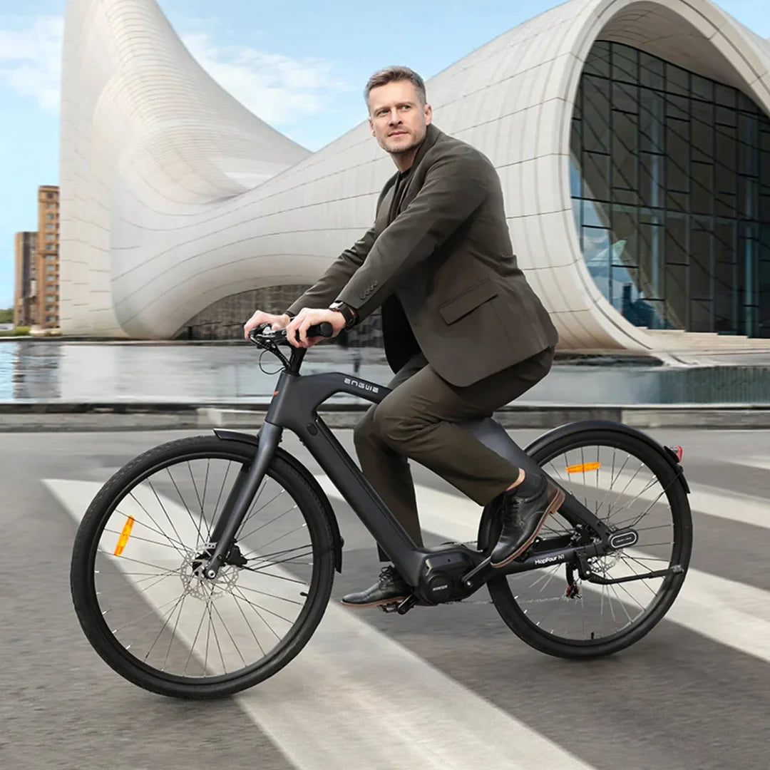 Izdelek na sliki je električno kolo, znamke Engwe, model Mapfour N1 Pro. Na sliki je moški v poslovnem obleku, ki se vozi na črnem električnem kolesu, medtem ko se v ozadju vidi moderna arhitektura in vodna površina. Kolo ima široke pnevmatike in elegantno obliko, kar poudarja njegovo urbano naravo.