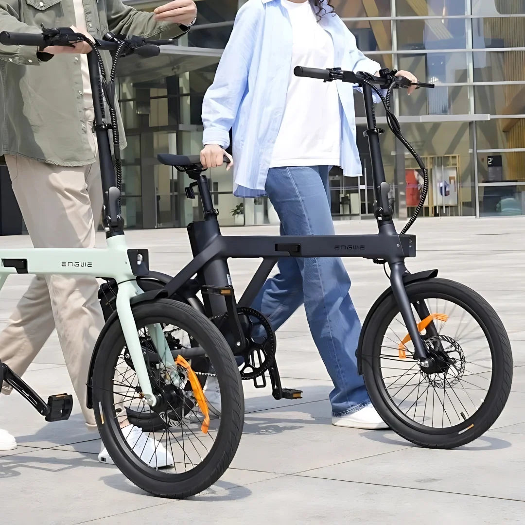 Na sliki sta prikazana dva električna kolesa znamke Engwe, model P20. Kolesi imata kompaktno zasnovo in sta opremljeni s širokimi pnevmatikami ter disk zavornim sistemom. Osebi, ki ju držita, sta oblečeni v sproščena oblačila, kar nakazuje na udobno vožnjo. Ozadje slike prikazuje moderno urbano okolje, kar dodatno poudarja praktičnost teh električnih koles.