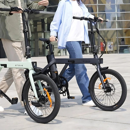 Na sliki sta prikazana dva električna kolesa znamke Engwe, model P20. Kolesi imata kompaktno zasnovo in sta opremljeni s širokimi pnevmatikami ter disk zavornim sistemom. Osebi, ki ju držita, sta oblečeni v sproščena oblačila, kar nakazuje na udobno vožnjo. Ozadje slike prikazuje moderno urbano okolje, kar dodatno poudarja praktičnost teh električnih koles.