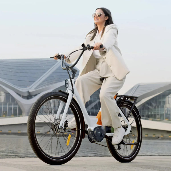 Izdelek je električno kolo znamke Engwe, model P275 ST. Na sliki je prikazana ženska, ki se vozi s kolesom ob vodi, oblečena v svetlo obleko. Kolo ima široke pnevmatike in moderno zasnovo, kar nakazuje na udobno vožnjo. V ozadju so vidne sodobne arhitekturne strukture.