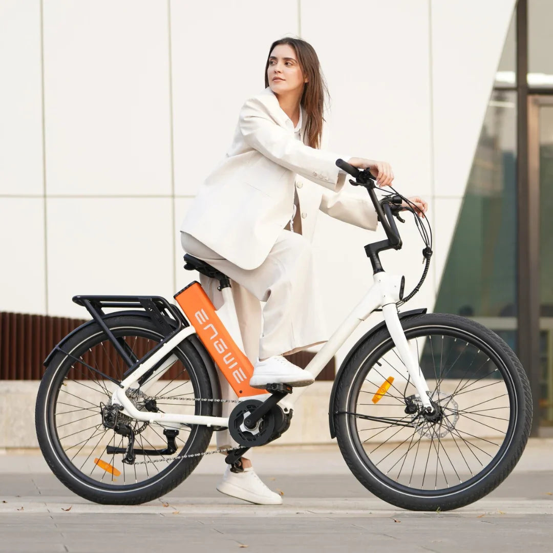 Izdelek je električno kolo znamke Engwe, model P275 ST. Na sliki je vidna ženska, ki sedi na kolesu, oblečena v svetlo obleko. Kolo je belo in ima značilno oranžno baterijo na okvirju ter nosilec za prtljago. V ozadju je moderna arhitektura, kar daje sliki sodoben videz.