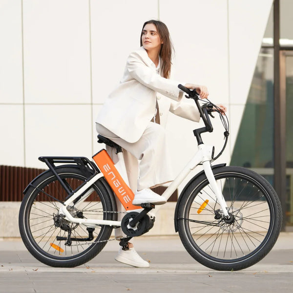 Izdelek je električno kolo znamke Engwe, model P275 ST. Na sliki je vidna ženska, ki sedi na kolesu, oblečena v svetlo obleko. Kolo je belo in ima značilno oranžno baterijo na okvirju ter nosilec za prtljago. V ozadju je moderna arhitektura, kar daje sliki sodoben videz.