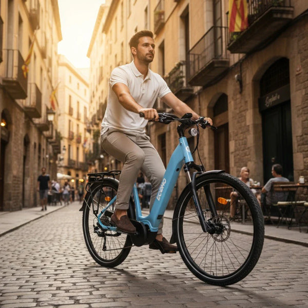 Na sliki je električno kolo znamke Econo, model Mare City EEC4LK. Kolo je svetlo modre barve in ima udoben okvir, kar omogoča enostavno vstopanje. Na sliki je moški, ki vozi kolo po slikoviti ulici s tlakovanjem, obdan z zgodovinskimi zgradbami in ljudmi, ki uživajo v okolici. Kolo je opremljeno z zadnjim prtljažnikom in ima dodatno opremo, kar ga naredi primernega za mestno vožnjo.