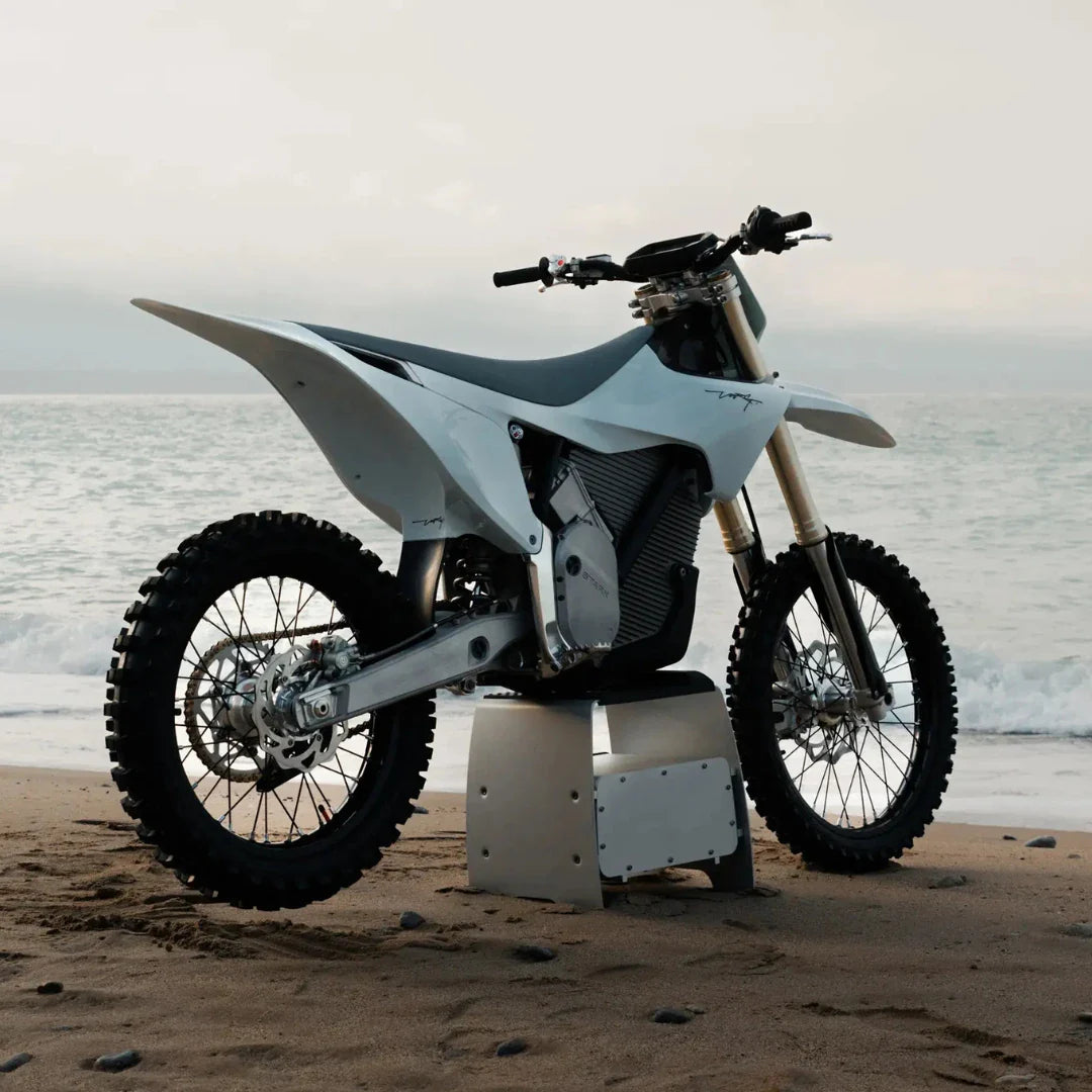 Električni motokros motocikel Stark Varg parkiran na peščeni plaži z oceanskimi valovi v ozadju.