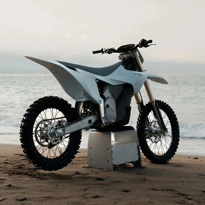 Električni motokros motocikel Stark Varg parkiran na peščeni plaži z oceanskimi valovi v ozadju.