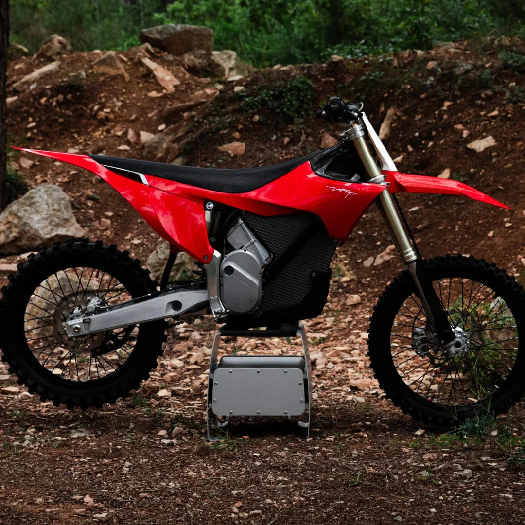Električni motokros Stark Varg v rdeči barvi, parkiran na skalnatem terenu z zelenjem v ozadju.