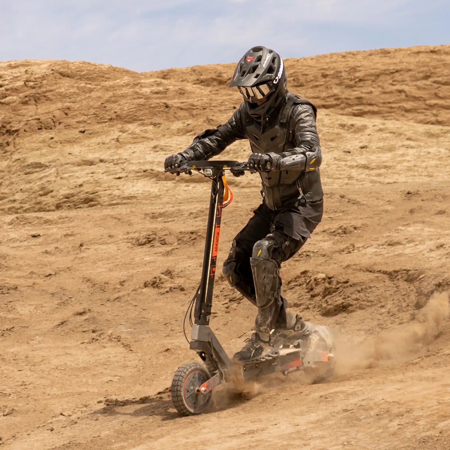 The product in the picture is an electric scooter, model Kukirin G2 Ultra. A person wearing protective gear is seen riding the scooter on uneven terrain. Dust is rising around them, indicating a dynamic ride.