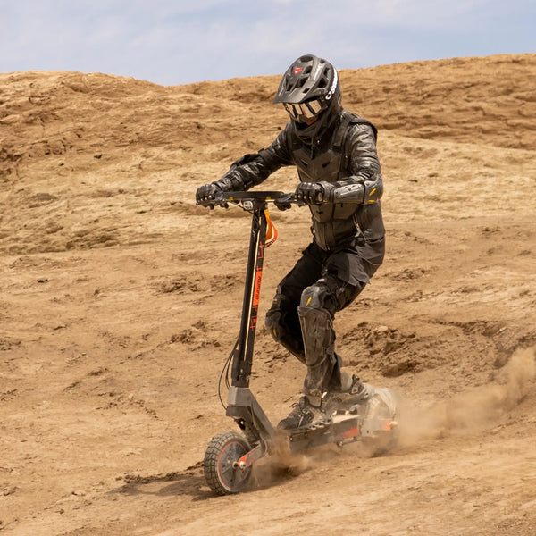 Izdelek na sliki je električni skiro, model Kukirin G2 Ultra. Na sliki je vidna oseba, oblečena v zaščitno opremo, ki vozi skiro po neravnem terenu. Okoli njega se dviga prah, kar nakazuje na dinamično vožnjo.