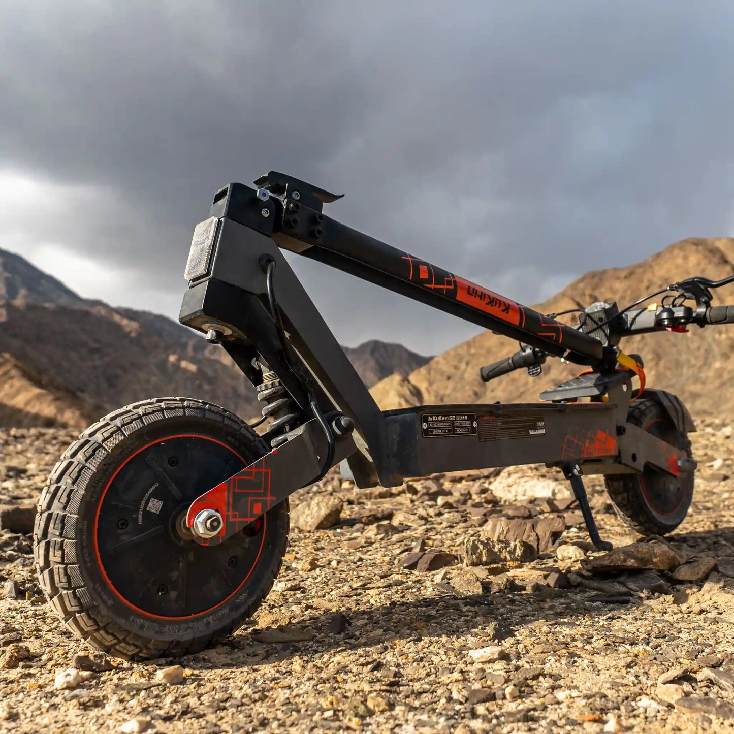 The picture shows a Kukirin electric scooter, model G2 Ultra. The scooter's robust construction is visible, featuring wide tires for better stability on terrain. The rocky landscape in the background suggests that the scooter is suitable for riding on more demanding terrains.