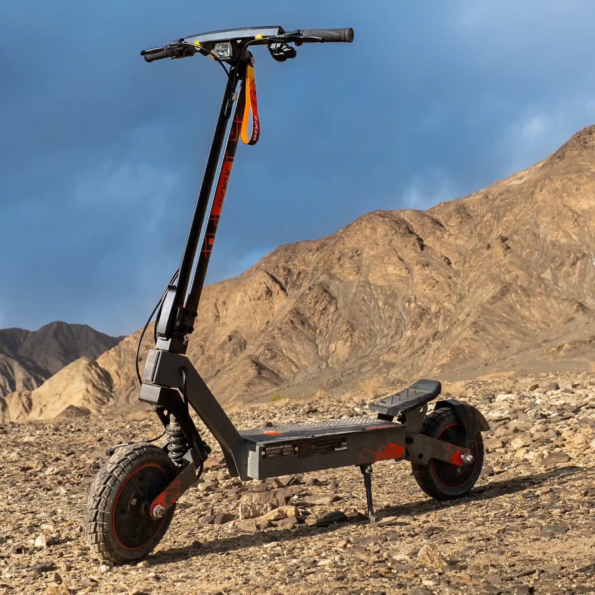 Izdelek na sliki je električni skiro, model Kukirin G2 Ultra. Na sliki je vidna robustna zasnova skiroja, ki je postavljen na kamenitem terenu, kar nakazuje njegovo primernost za vožnjo po zahtevnejših površinah. Ima široka kolesa in vzmetenje, kar prispeva k udobni vožnji. Rdeči detajli na črni strukturi dodajajo estetski pridih.