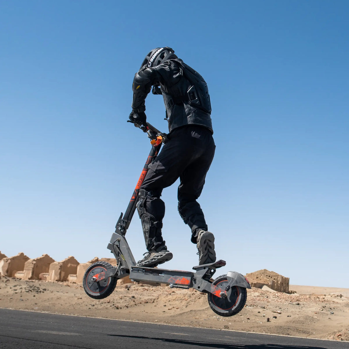 Na sliki je električni skiro, model Kukirin G2 Ultra. Vidimo voznika, ki skoči s skiroja na odprtem terenu, oblečen v zaščitno opremo, kar nakazuje na športno uporabo. V ozadju so vidne peščene strukture, kar daje občutek avanture in prostega prostora.