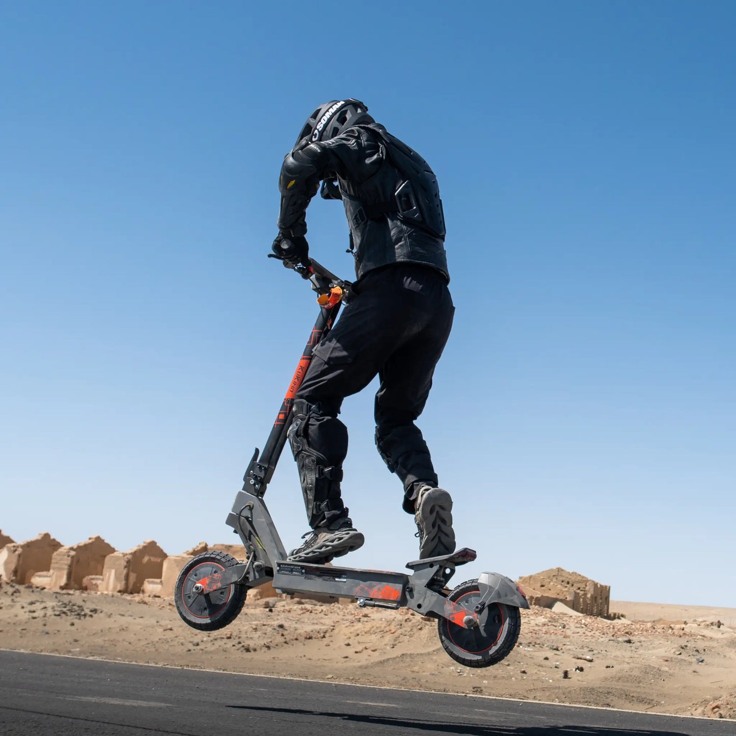 The image shows an electric scooter, the Kukirin G2 Ultra model. We see a rider, dressed in protective gear, jumping with the scooter in an open terrain, which suggests sporty use. In the background, sandy structures are visible, giving a sense of adventure and open space.