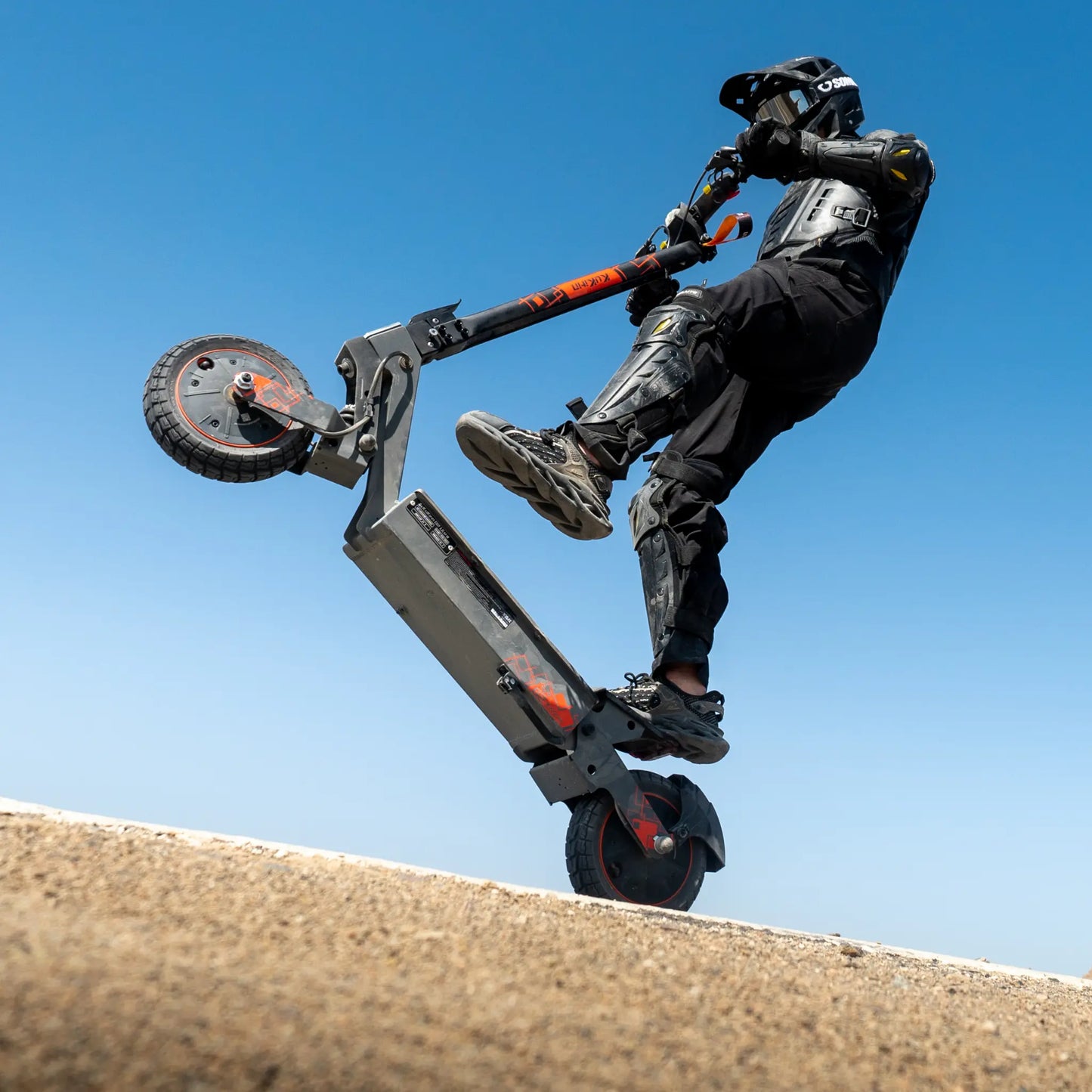 The image shows an electric scooter, the Kukirin G2 Ultra model. We see a rider performing a trick, lifting the scooter onto its rear wheel. He is wearing protective gear, which indicates a sporty ride. The background is a clear blue sky, giving the image a dynamic look.
