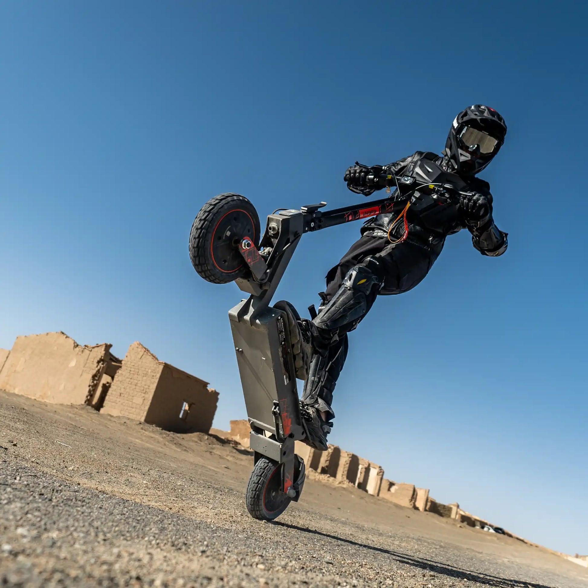 Na sliki je električni skiro, model Kukirin G2 Ultra. Vidimo voznika, oblečenega v zaščitno opremo, ki izvaja trik z dvigovanjem sprednjega kolesa. Ozadje prikazuje ruševine, kar daje sliki pustolovski občutek. Skuter ima robustno zasnovo in velike pnevmatike, kar nakazuje na njegovo zmogljivost na različnih terenih.