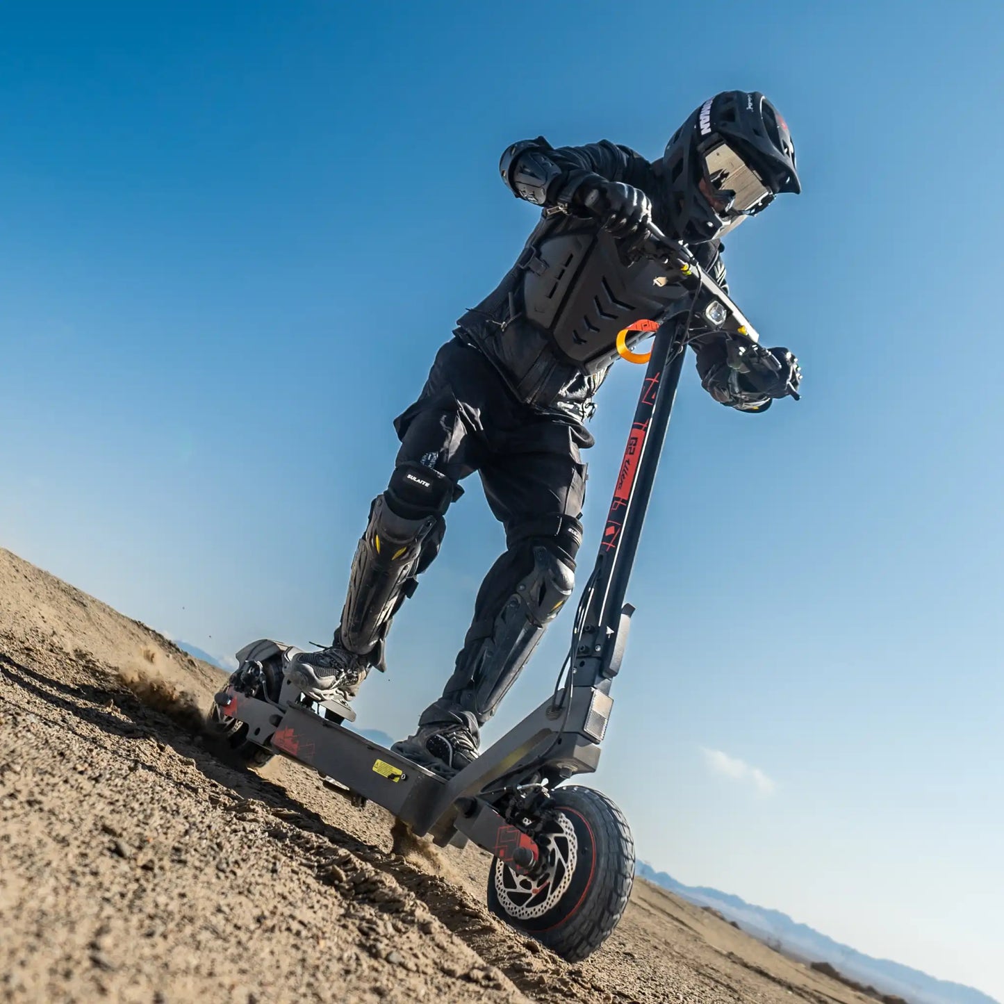 The product in the picture is an electric scooter, model Kukirin G2 Ultra. The picture shows a rider, dressed in protective gear, riding on a sandy surface. The scooter has a robust design and wide tires, which indicates its ability to ride on various terrains. The background shows a clear blue sky and mountain outlines.