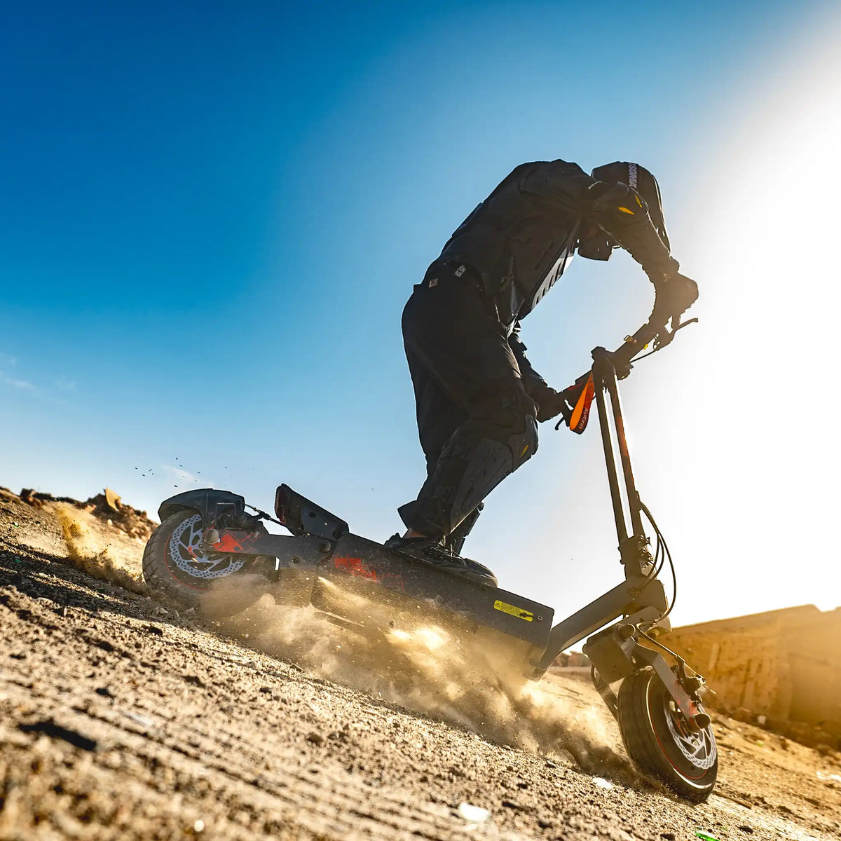 Na sliki je električni skiro znamke Kukirin, model G2 Ultra. Vidimo voznika, ki se vozi po neurejeni površini, kar povzroča prah in kamenčke, ki se dvigajo okoli. Voznik je oblečen v zaščitno opremo, kar nakazuje na športno vožnjo in avanturistični slog. Ozadje je jasno in sončno, kar prispeva k dinamični atmosferi slike.