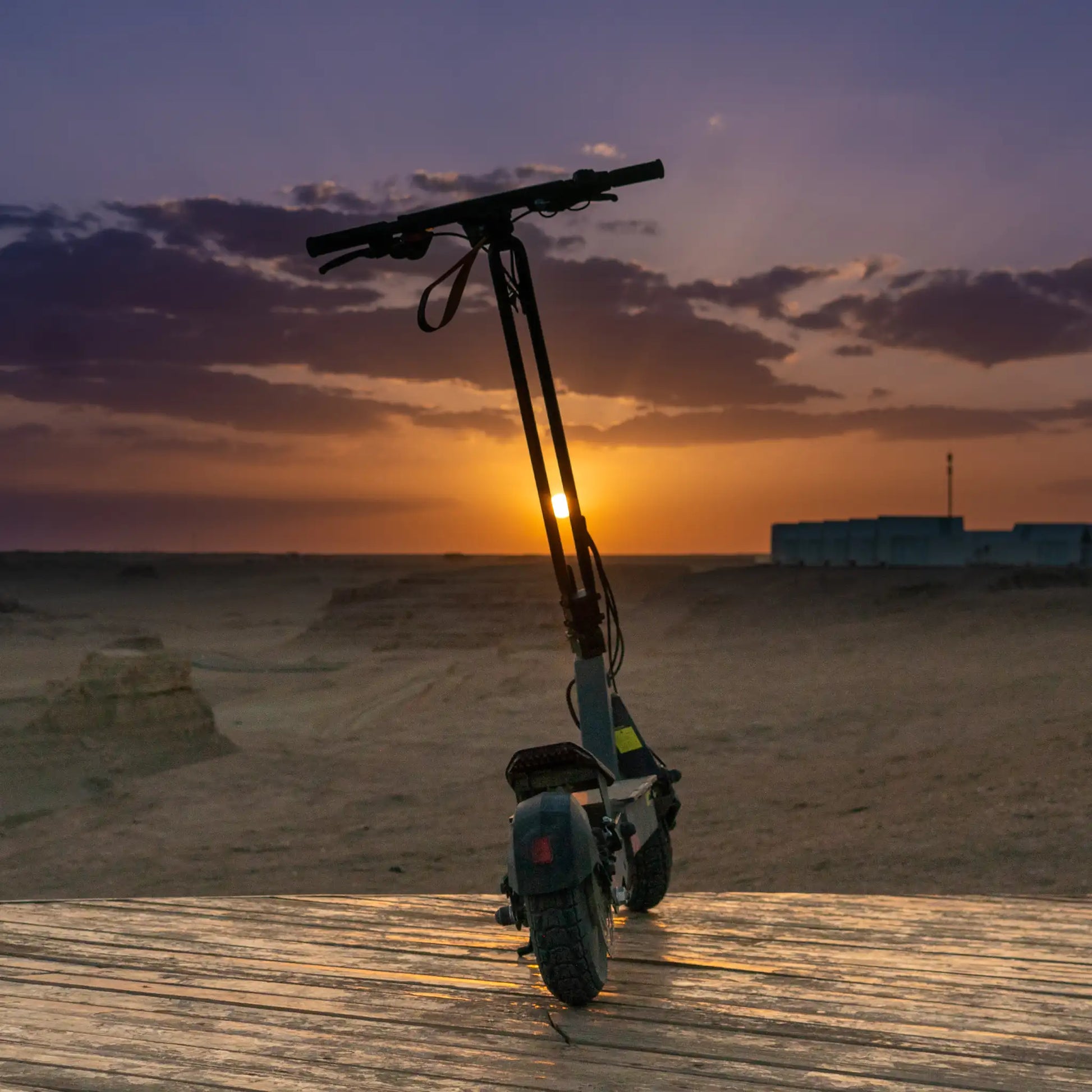 Na sliki je električni skiro znamke Kukirin, model G2 Ultra. Izdelek je postavljen na leseno površino, v ozadju pa je vidna pokrajina ob sončnem zahodu. Skrbno oblikovan skiro ima široka kolesa, kar nakazuje na stabilnost in udobno vožnjo. Atmosfera slike je mirna in naravna, kar dodatno poudarja funkcionalnost skiroja v prostem okolju.