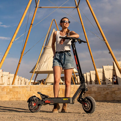 Na sliki je električni skiro znamke Kukirin, model G2 Ultra. Vidna je ženska, ki se naslanja na skiro, oblečena v kratke hlače in majico, ter nosi sončna očala. V ozadju so vidne strukture, ki spominjajo na šotore, kar daje sliki sproščen in poletni občutek.