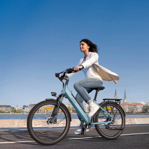 Na sliki je električno kolo znamke Engwe, model P275 SE. Prikazuje žensko, ki vozi kolo ob reki, oblečeno v svetlo jakno in kavbojke. Kolo ima modro barvo in je opremljeno s standardnimi komponentami, kot so krmilo in sedež, ter je v slikovitem okolju z mestno arhitekturo v ozadju.