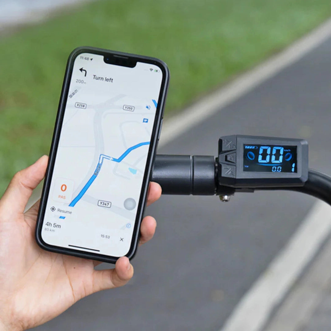 Izdelek na sliki je električno kolo, model Engwe P275 SE. Na sliki je prikazana roka, ki drži pametni telefon z navigacijsko aplikacijo, medtem ko je na krmilu kolesa vidna digitalna merilna enota, ki prikazuje hitrost in druge podatke. V ozadju je vidna pot, kar nakazuje na uporabo kolesa v naravnem okolju.