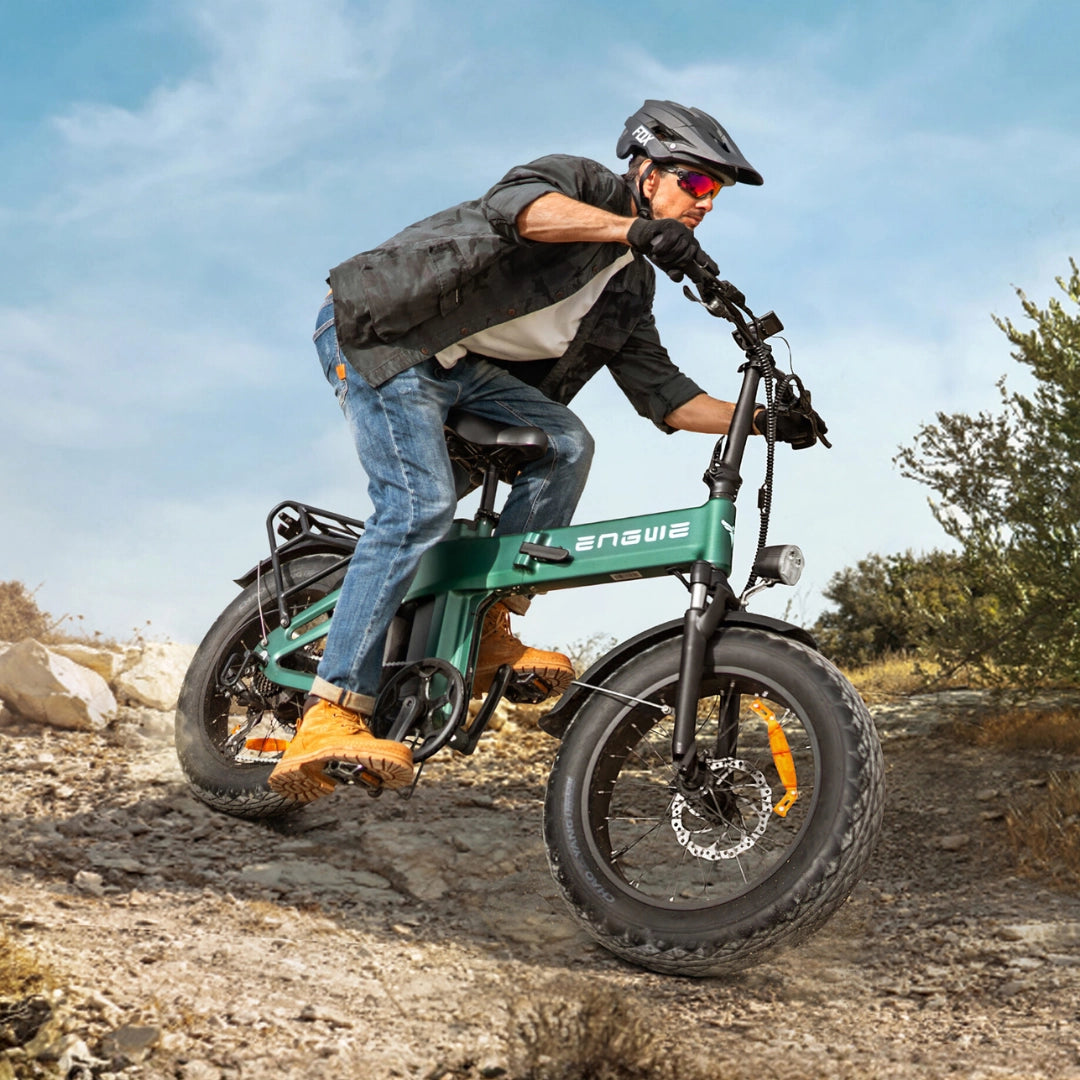 Na sliki je električno kolo znamke Engwe, model EP-2 3.0 Boost. Kolo je prikazano med vožnjo po neravnem terenu, kar nakazuje na njegovo zmogljivost in primernost za avanturistično vožnjo. Kolesar nosi zaščitno čelado in rokavice, kar poudarja varnost pri vožnji.