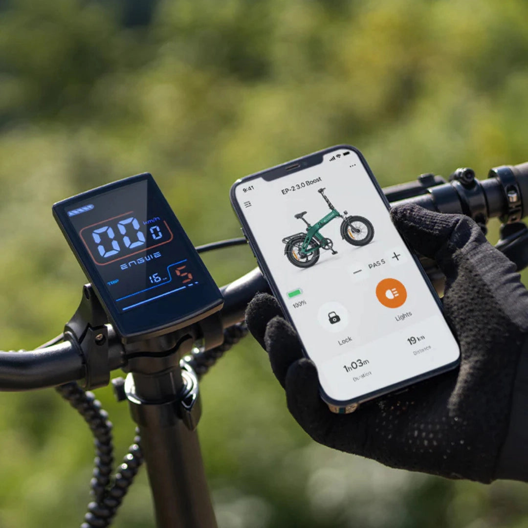 Na sliki je električno kolo znamke Engwe, model EP-2 3.0 Boost. Vidna je digitalna zaslon na krmilu, ki prikazuje hitrost in druge podatke, ter pametni telefon, ki prikazuje informacije o kolesu, vključno z nivojem baterije in različnimi nastavitvami. Kolo je zeleno in ima zložljivo zasnovo.