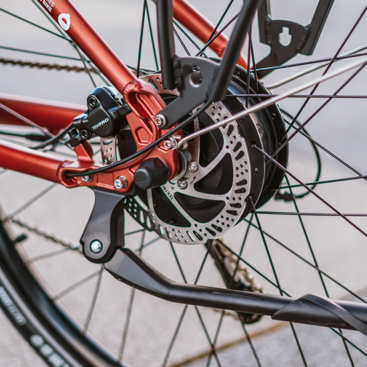 Izdelek je električno kolo, model Econo Apes EER4. Na sliki je prikazan zadnji del kolesa, kjer je vidna električna motorna enota, disk zavora in del okvirja v rdeči barvi. Kolo ima tudi vidne komponente, kot so kolesni obročki in veriga, kar nakazuje na robustno zasnovo za urbano vožnjo.