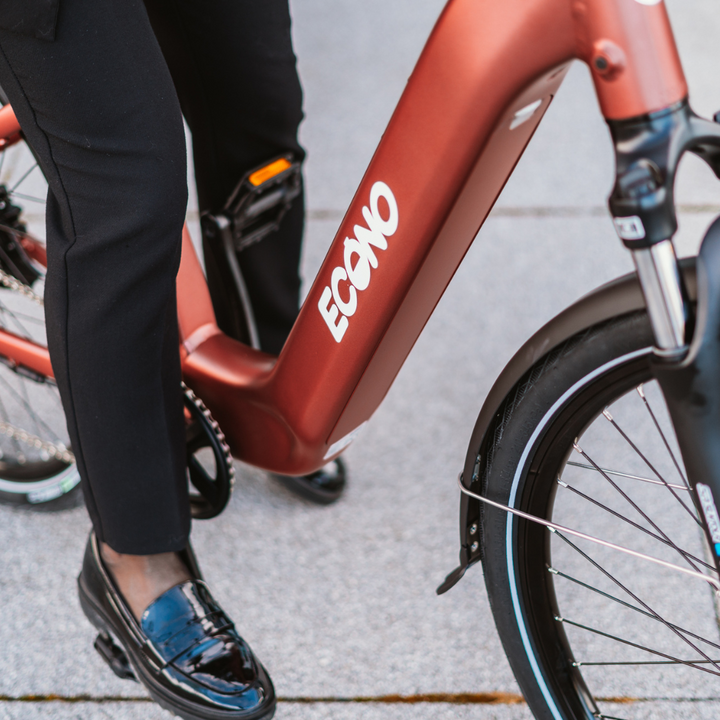 Na sliki je električno kolo znamke Econo, model EER4. Vidna je oseba, ki stoji ob kolesu, pri čemer je poudarjen okvir v rjavi barvi z logotipom Econo. Kolo ima klasične kolesarske komponente, vključno s sprednjim kolesom in pedalom. Oseba nosi elegantna oblačila, kar nakazuje na urbano uporabo kolesa.