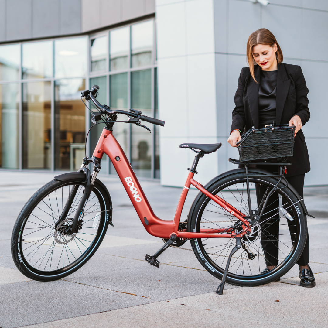 Na sliki je električno kolo znamke Econo, model EER4. Kolo ima sodoben dizajn in je v rdeči barvi. Na sliki je tudi ženska, ki stoji ob kolesu in drži košaro, kar nakazuje na praktičnost in uporabnost kolesa za mestno vožnjo. Ozadje je urbano, kar dodatno poudarja namen izdelka.