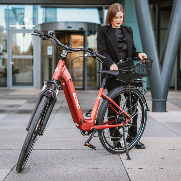 Na sliki je električno kolo znamke Econo, model EER4. Kolo je rdeče barve in ima sprednji koš za shranjevanje. Na sliki je vidna ženska oseba, ki stoji ob kolesu in se pripravlja, da nekaj vzame iz koša. Ozadje prikazuje moderno stavbo z velikimi steklenimi površinami.
