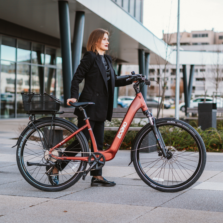 Na sliki je električno kolo znamke Econo, model EER4. Kolo ima moderno obliko in je opremljeno s košarico na zadnjem delu. Oseba, ki stoji ob kolesu, nosi elegantno obleko, kar nakazuje, da je kolo primerno za mestno vožnjo. Ozadje prikazuje sodobno urbano okolje z arhitekturo in parkiriščem.