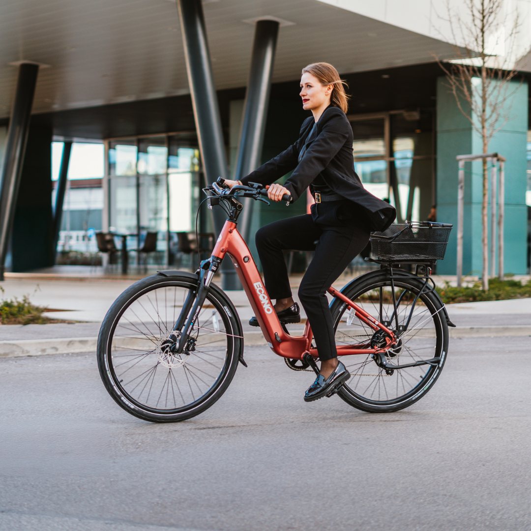 Izdelek je električno kolo znamke Econo, model EER4. Na sliki je vidna ženska, oblečena v poslovno obleko, ki vozi električno kolo po urbanem okolju. Kolo ima rdečo barvo in je opremljeno s košarico na zadnjem delu, kar nakazuje na praktičnost in udobje pri vožnji. Ozadje prikazuje sodobno arhitekturo in urejeno okolje.