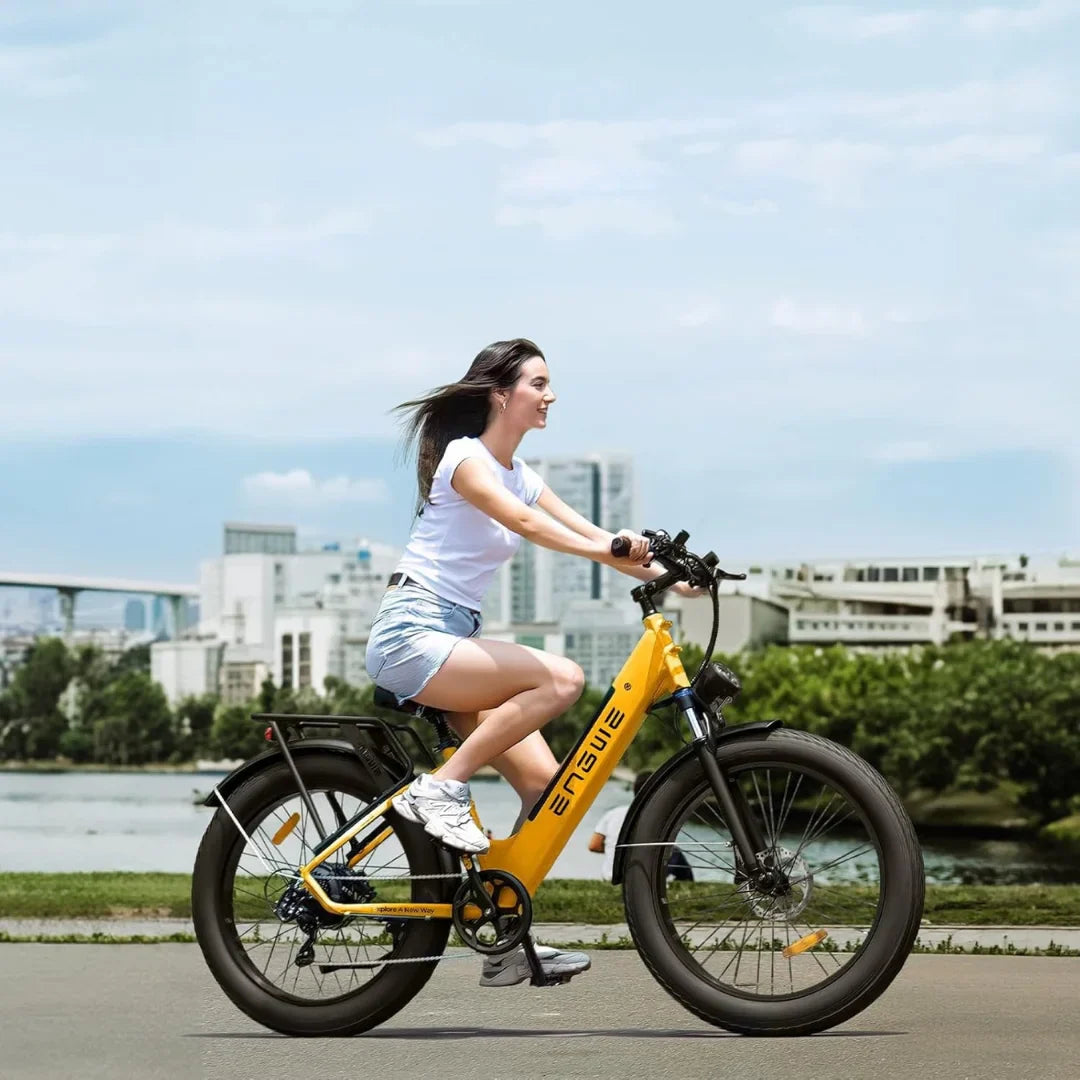 Na sliki je električno kolo znamke Engwe, model E26. Kolo je v svetlo rumeni barvi in ima široke pnevmatike, kar daje občutek stabilnosti. Na njem se vozi mlada ženska, oblečena v belo majico in kratke hlače, medtem ko se vozi po urejeni poti ob vodi. V ozadju so vidne moderne zgradbe in zelenje, kar ustvarja prijetno urbano okolje.