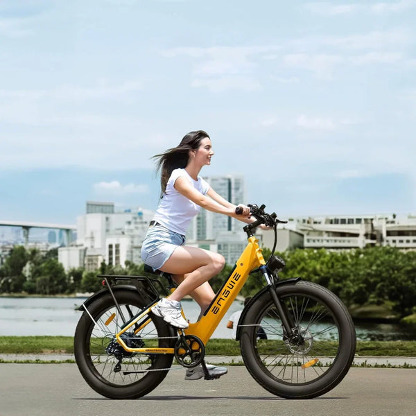 Na sliki je električno kolo znamke Engwe, model E26. Kolo je v svetlo rumeni barvi in ima široke pnevmatike, kar daje občutek stabilnosti. Na njem se vozi mlada ženska, oblečena v belo majico in kratke hlače, medtem ko se vozi po urejeni poti ob vodi. V ozadju so vidne moderne zgradbe in zelenje, kar ustvarja prijetno urbano okolje.