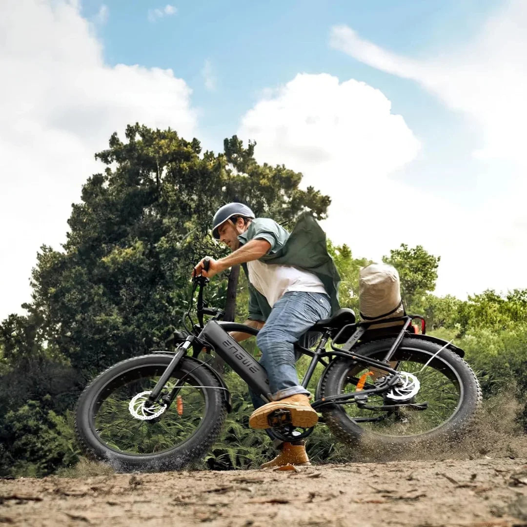 Na sliki je električno kolo znamke Engwe, model E26. Prikazuje moškega, ki vozi kolo po neurejeni površini, kar nakazuje na njegovo zmogljivost v terenu. Kolo ima široke pnevmatike in je opremljeno s prtljažnikom, na katerem je nahrbtnik. V ozadju so drevesa in modro nebo, kar ustvarja naravno okolje za vožnjo.