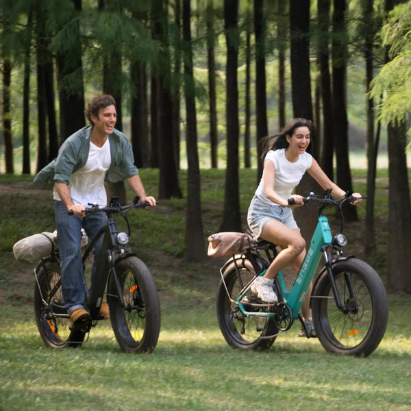 Izdelek na sliki je električno kolo, znamke Engwe, model E26. Na sliki sta prikazana moški in ženska, ki se zabavata med vožnjo po naravi. Obe kolesi imata široke pnevmatike in udobne sedeže, kar nakazuje na sproščeno in prijetno vožnjo. Okolica je gozdnata, kar ustvarja prijetno in naravno vzdušje.