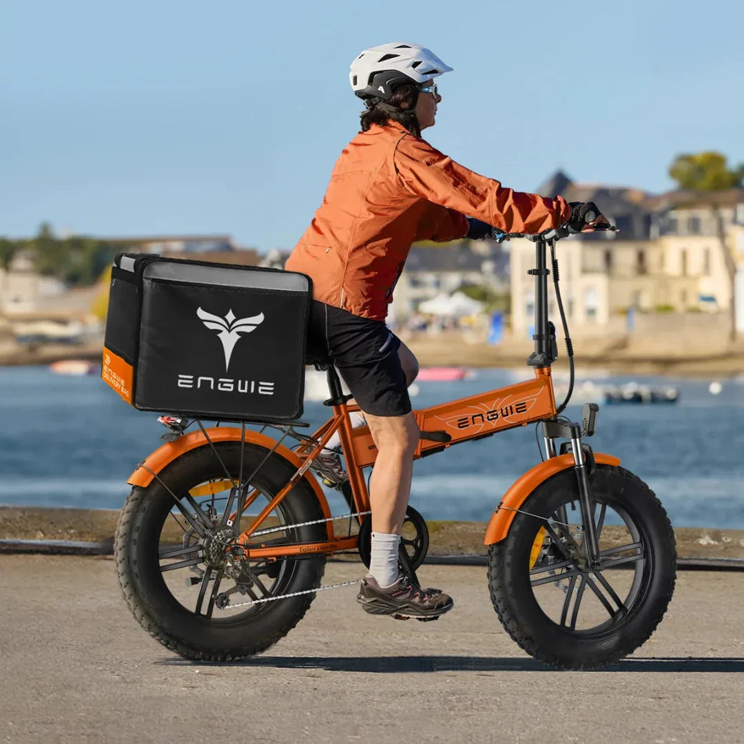 Izdelek na sliki je električno kolo znamke Engwe, model EP-2 Boost. Na sliki je vidna oseba, ki vozi kolo ob vodi, oblečena v oranžno jakno in čelado. Kolo ima široke pnevmatike in na zadnjem delu je pritrjen črn nahrbtnik z logotipom Engwe, kar nakazuje na možnost dostave ali prevoza.