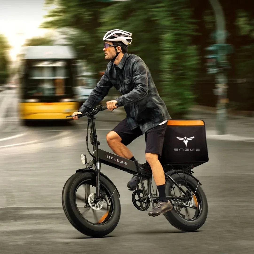 Izdelek na sliki je električno kolo znamke Engwe, model EP-2 Boost. Na sliki je prikazan moški, ki vozi kolo po mestni ulici. Kolo ima široke pnevmatike, kar omogoča boljšo stabilnost, in je opremljeno s prtljažnikom, ki je namenjen prevozu stvari. Moški nosi čelado in športna oblačila, kar nakazuje na aktivno vožnjo. V ozadju so vidni mestni elementi, kar dodatno poudarja urbano okolje.