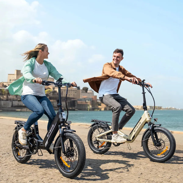 Izdelek na sliki je električno kolo znamke Engwe, model L20. Na sliki sta dva kolesarja, moški in ženska, ki se vozita ob obali. Obe kolesi imata široke gume in moderni dizajn, ženska nosi svetlo zeleni plašč, moški pa rjavo jakno. V ozadju je vidna obala in modro nebo.