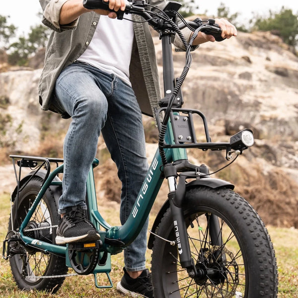 Na sliki je električno kolo znamke Engwe, model L20 Boost. Kolo ima široke pnevmatike, kar omogoča boljšo stabilnost in udobje pri vožnji po različnih terenih. Oseba na kolesu nosi športna oblačila in se nahaja na naravni lokaciji z skalnatimi površinami v ozadju. Kolo je opremljeno z nosilcem in svetilko, kar nakazuje na njegovo praktičnost za mestno in terensko uporabo.