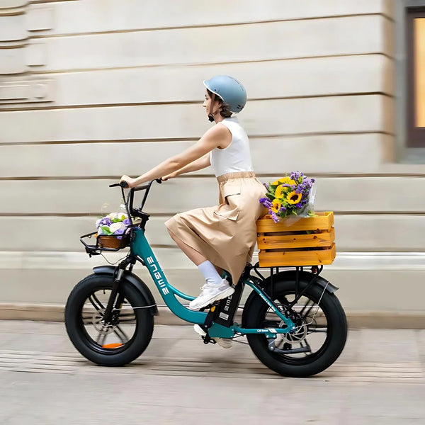 Na sliki je električno kolo znamke Engwe, model L20 Boost. Kolo ima široke pnevmatike in je opremljeno s košaro na zadnjem delu, v kateri so cvetlice. Na kolesu vozi ženska, ki nosi čelado in oblečena je v svetlo obleko. Ozadje prikazuje urbano okolje.