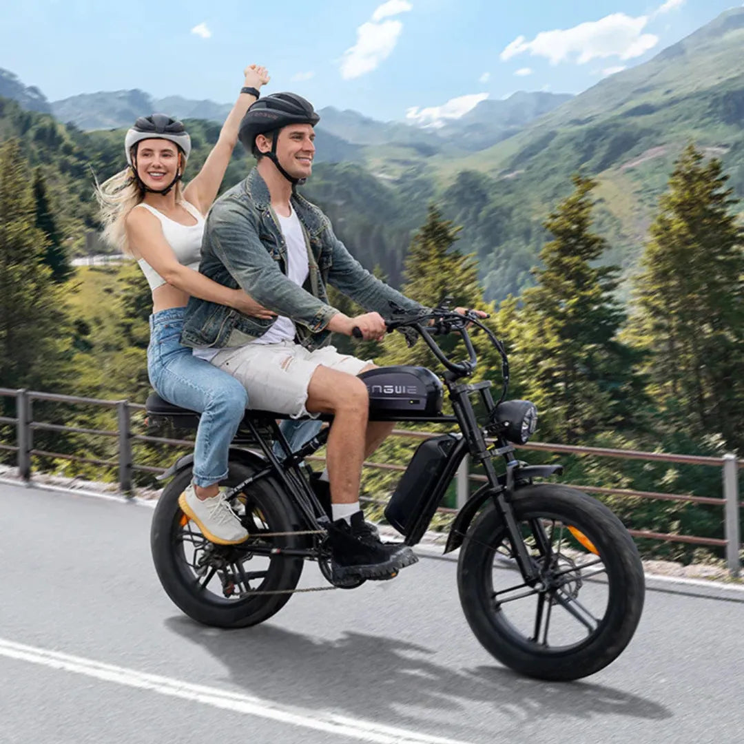 Izdelek na sliki je električno kolo znamke Engwe, model M1. Na sliki sta vidna dva človeka, ki se vozita na kolesu po slikoviti cesti ob gozdnatem območju. Oboževalca kolesarjenja sta oblečena v športna oblačila in nosita čelade. Kolo ima širše pnevmatike in moderno zasnovo, kar nakazuje na udobno vožnjo.