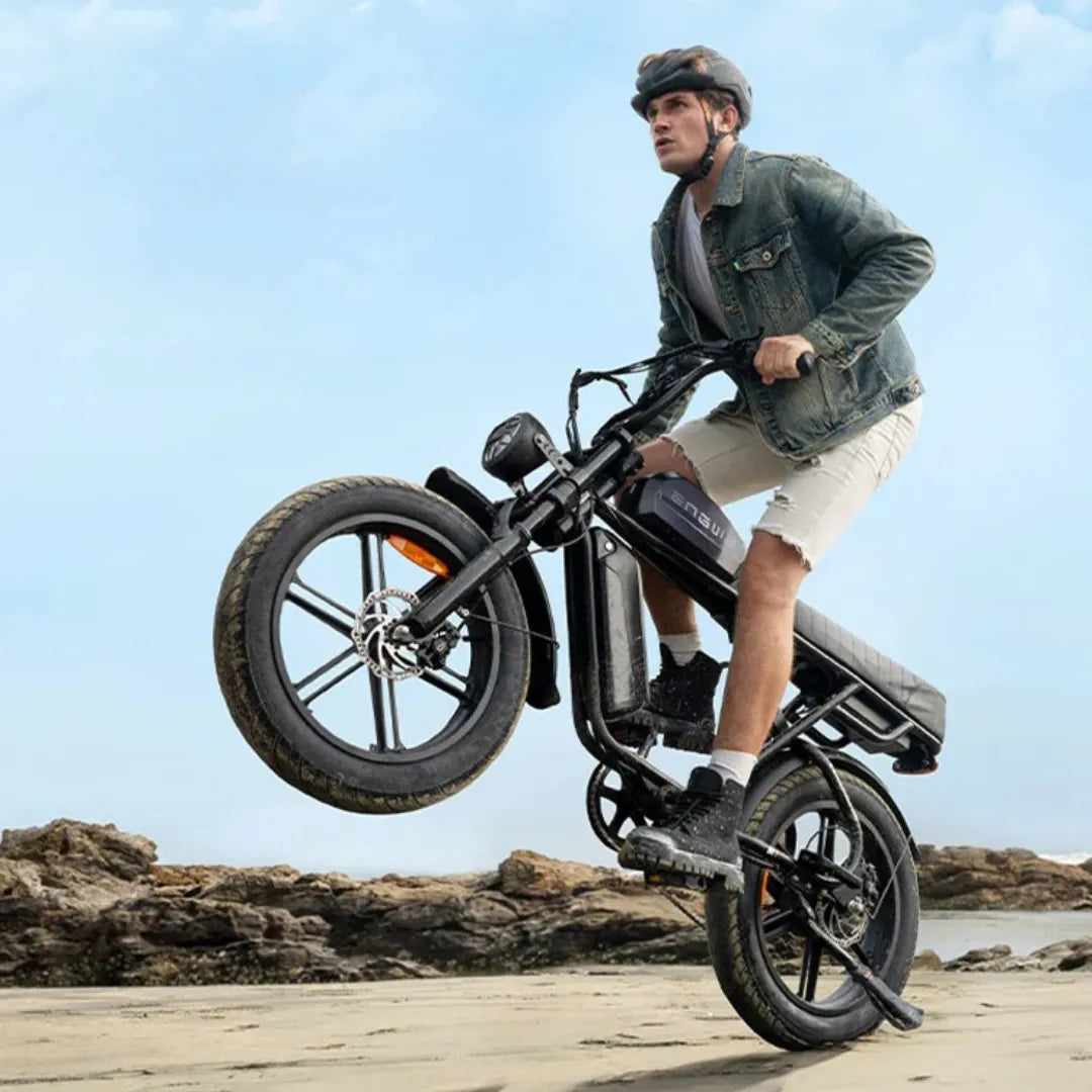 Na sliki je električno kolo znamke Engwe, model M1. Prikazuje moškega, ki izvaja trik z dvigovanjem sprednjega kolesa nad tlemi, medtem ko se vozi po plaži. Kolo ima robustno zasnovo, široka kolesa in je opremljeno z električnim motorjem ter baterijo, kar omogoča udobno vožnjo po različnih terenih. Moški nosi čelado in je oblečen v denim jakno ter kratke hlače.