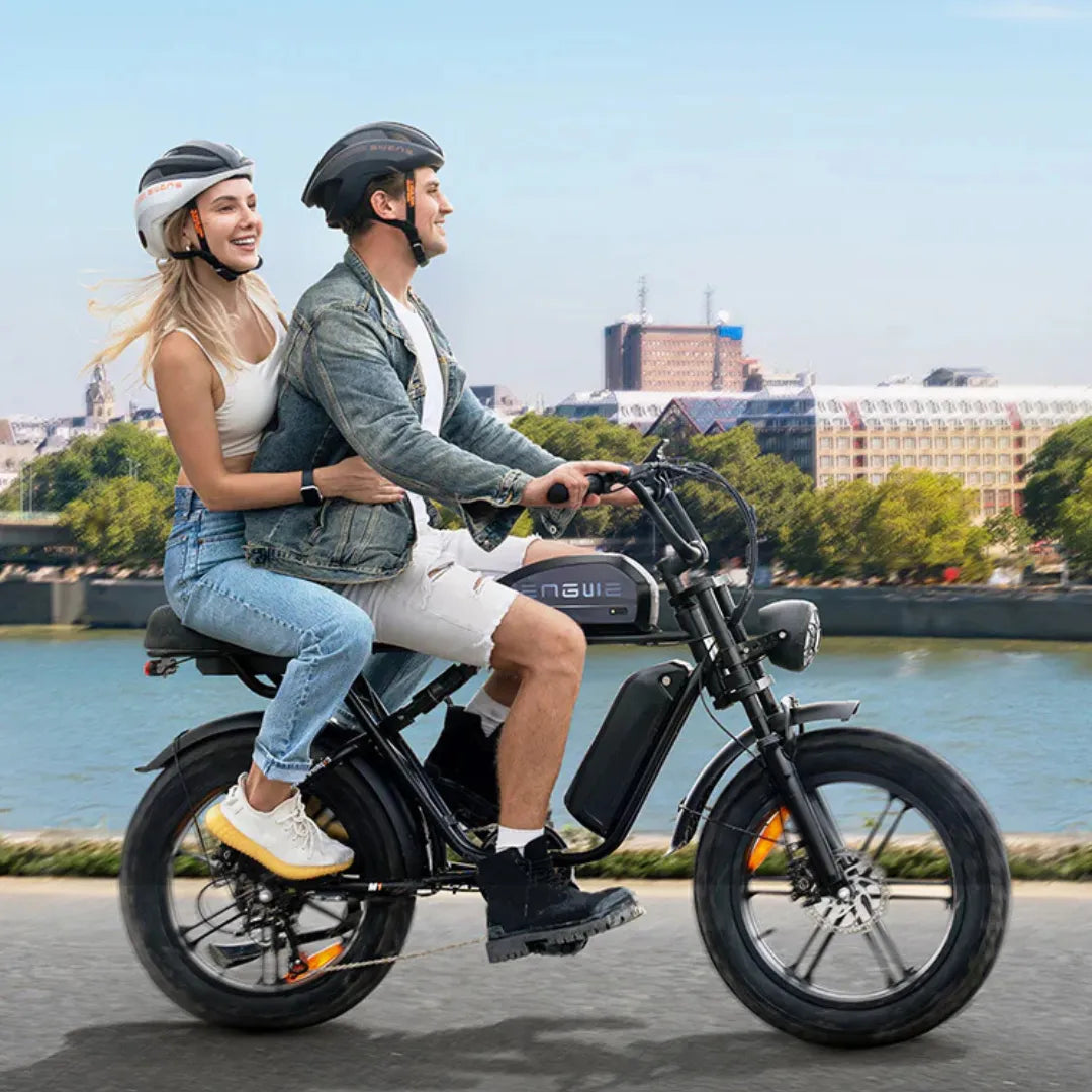 Izdelek na sliki je električno kolo, model Engwe M1. Na kolesu sta vidna dva voznika, moški in ženska, ki nosita čelade. Kolo ima široke pnevmatike in je zasnovano za udobno vožnjo. V ozadju je slikovit pogled na reko in mestno arhitekturo, kar ustvarja prijetno vzdušje.