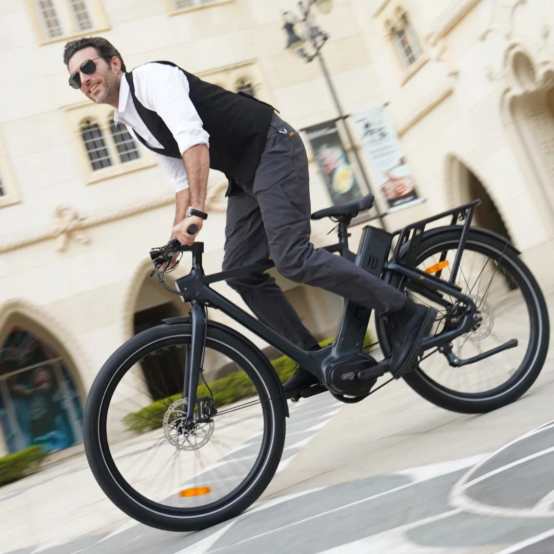 Izdelek je električno kolo znamke Engwe, model P275 Pro. Na sliki je moški, oblečen v elegantno obleko, ki se vozi na temnem električnem kolesu. Kolo ima široke pnevmatike in opremo za udobno vožnjo, vidna pa je tudi baterija na okvirju. Ozadje prikazuje arhitekturne elemente in mestno okolje.