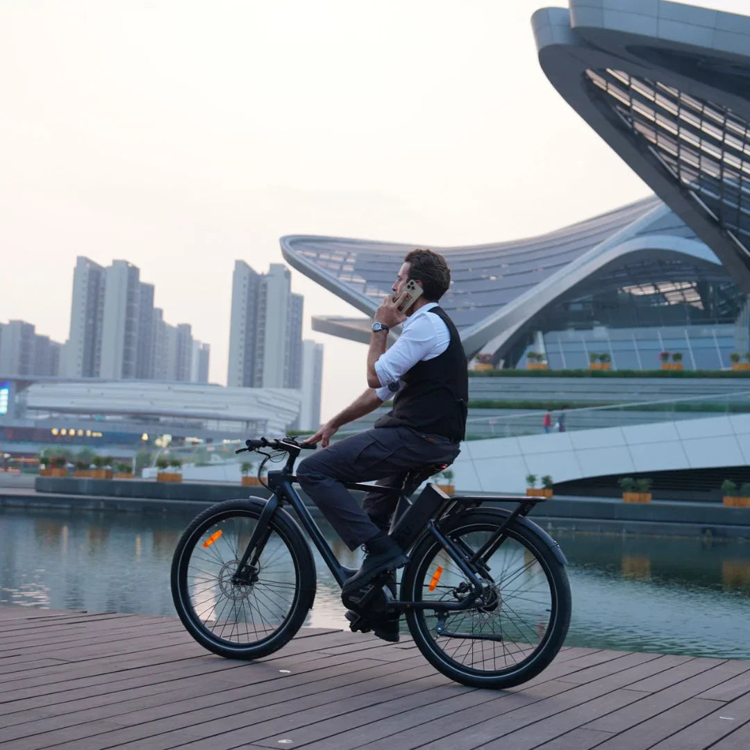 Na sliki je električno kolo znamke Engwe, model P275 Pro. Na kolesu sedi moški v formalni obleki in govori po telefonu. V ozadju je sodobna arhitektura in vodna površina, kar ustvarja urbani ambient. Kolo ima široke pnevmatike in je opremljeno z reflektorji za boljšo vidnost.