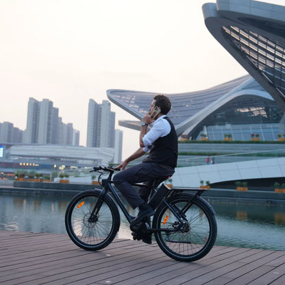 Na sliki je električno kolo znamke Engwe, model P275 Pro. Na kolesu sedi moški v formalni obleki in govori po telefonu. V ozadju je sodobna arhitektura in vodna površina, kar ustvarja urbani ambient. Kolo ima široke pnevmatike in je opremljeno z reflektorji za boljšo vidnost.