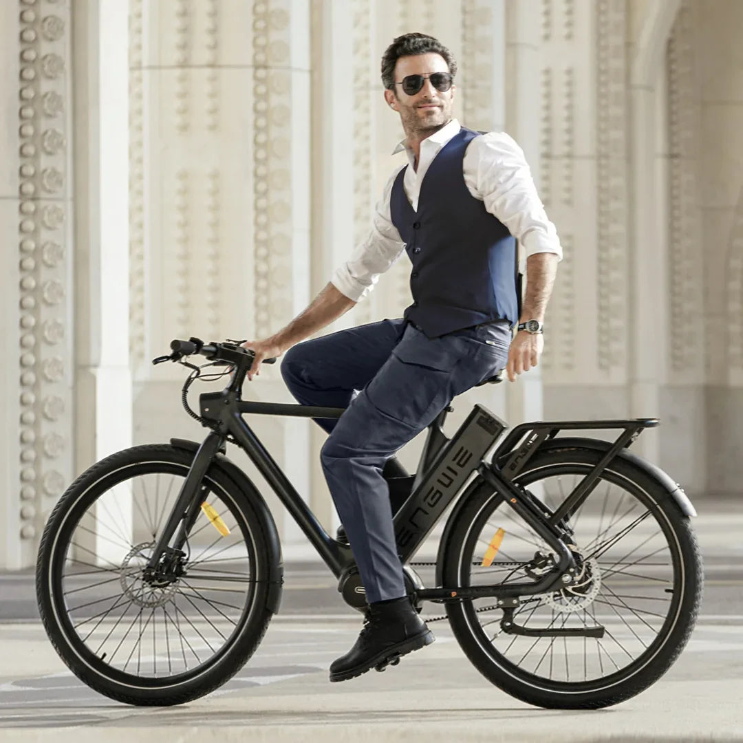 Na sliki je električno kolo znamke Engwe, model P275 Pro. Kolo je moderno zasnovano, s črnimi okvirji in širokimi pnevmatikami, kar omogoča udobno vožnjo. Na kolesu sedi moški v elegantni obleki, kar daje vtis, da je kolo primerno tako za prosti čas kot tudi za mestno vožnjo. V ozadju je arhitekturna struktura, ki dopolnjuje sodoben videz slike.