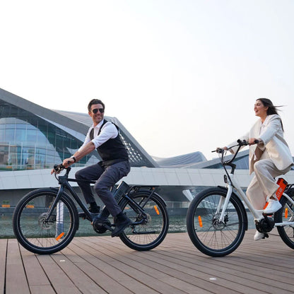 Izdelek na sliki je električno kolo, model Engwe P275 Pro. Na sliki sta vidna moški in ženska, ki se vozita na dveh električnih kolesih ob vodi, v ozadju pa je moderna arhitektura. Moški je oblečen v eleganten outfit, medtem ko ženska nosi svetlo obleko. Kolesi imata sodoben dizajn in sta opremljeni z električnimi komponentami.